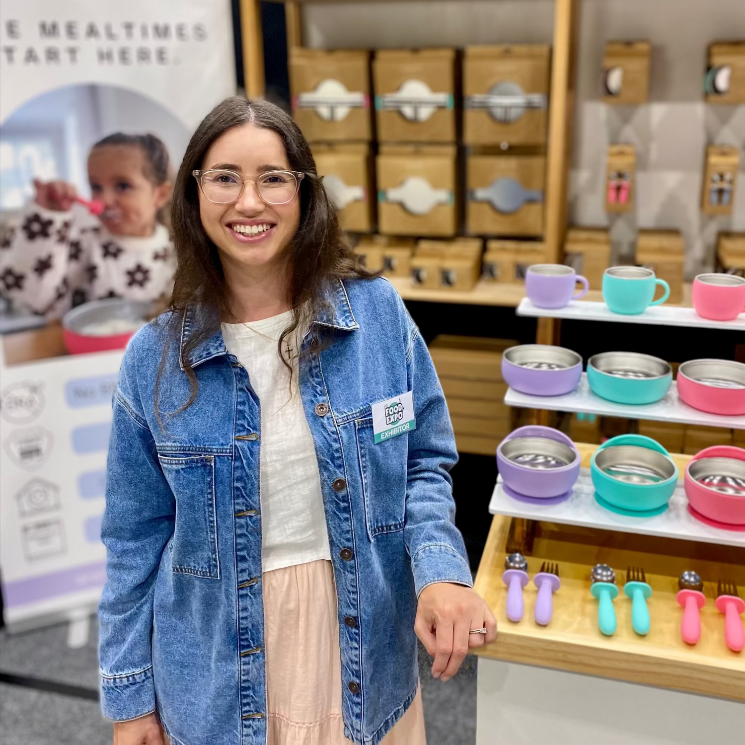 A smiling woman wearing a denim jacket and a light pink skirt stands in front of a NOMZY product display booth. The display features wooden storage boxes, colorful mugs, and bowls on shelves.